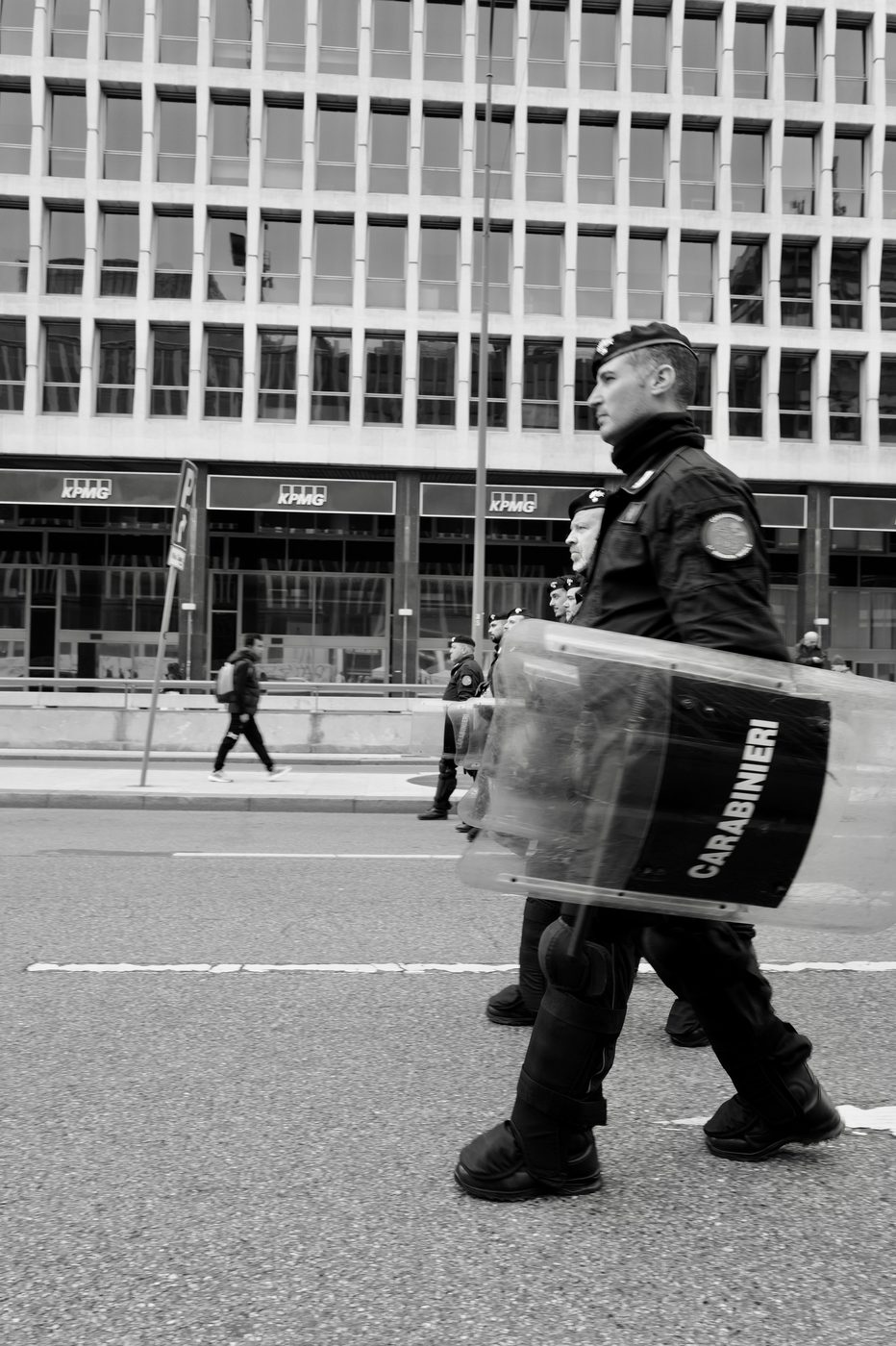 Carabiniere in riot gear, branded shield in hand, marching past an office building, Milan