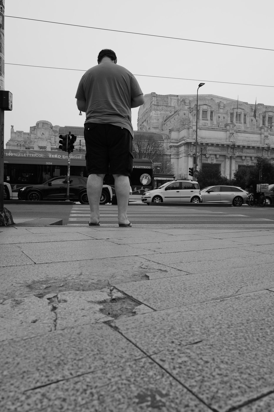 Man seen from behind waiting to cross, the monumental facade of the central station in the distance, Milan