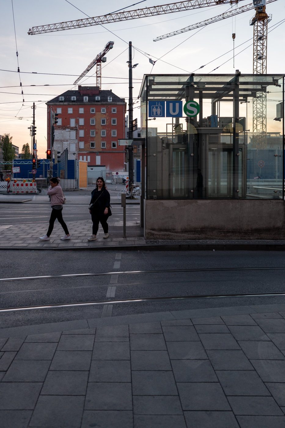 Two passers-by cross at dusk near a glazed tram entrance, Munich