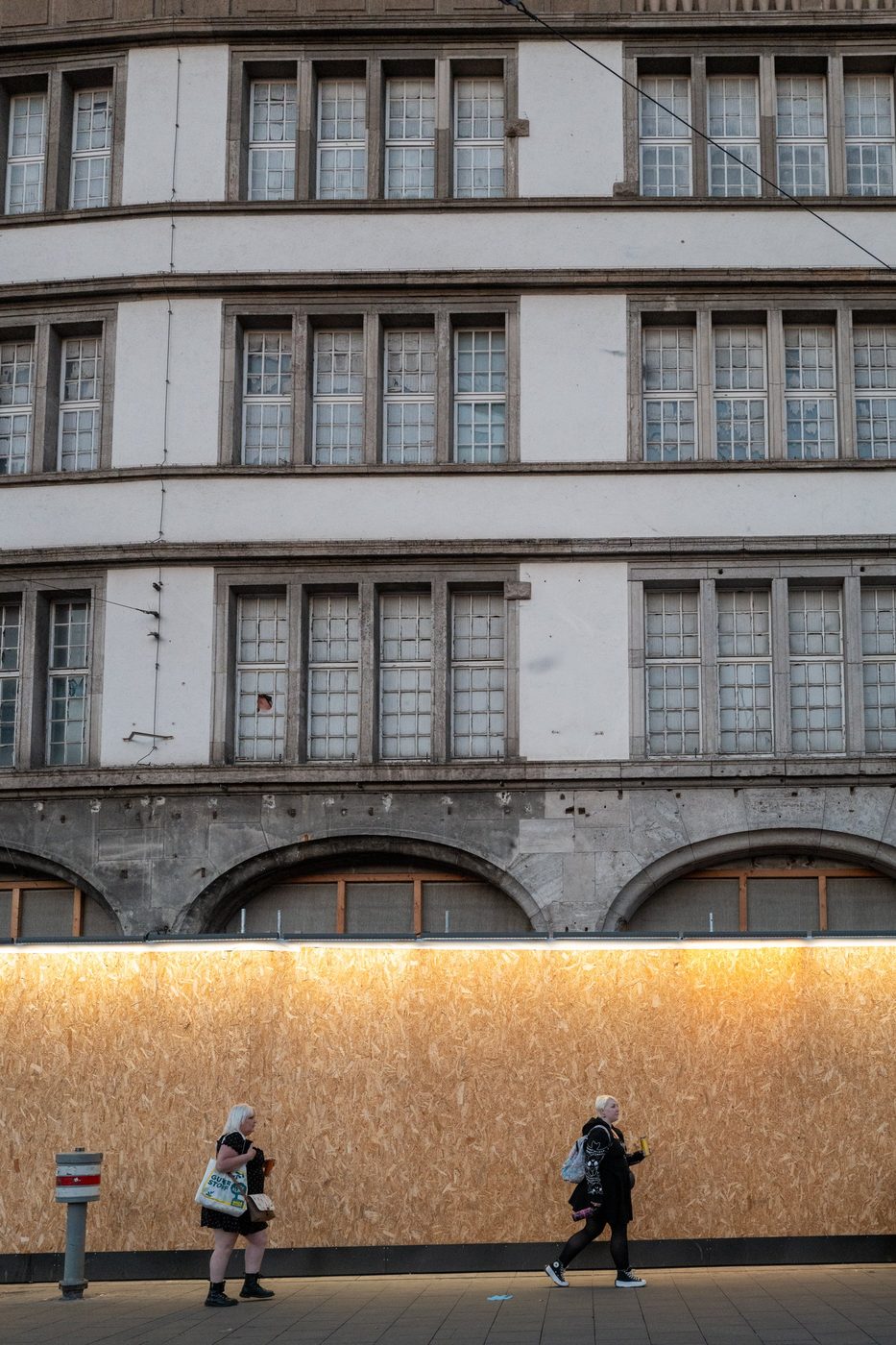 Two passers-by walk along a raw wooden wall in front of an old façade, Munich
