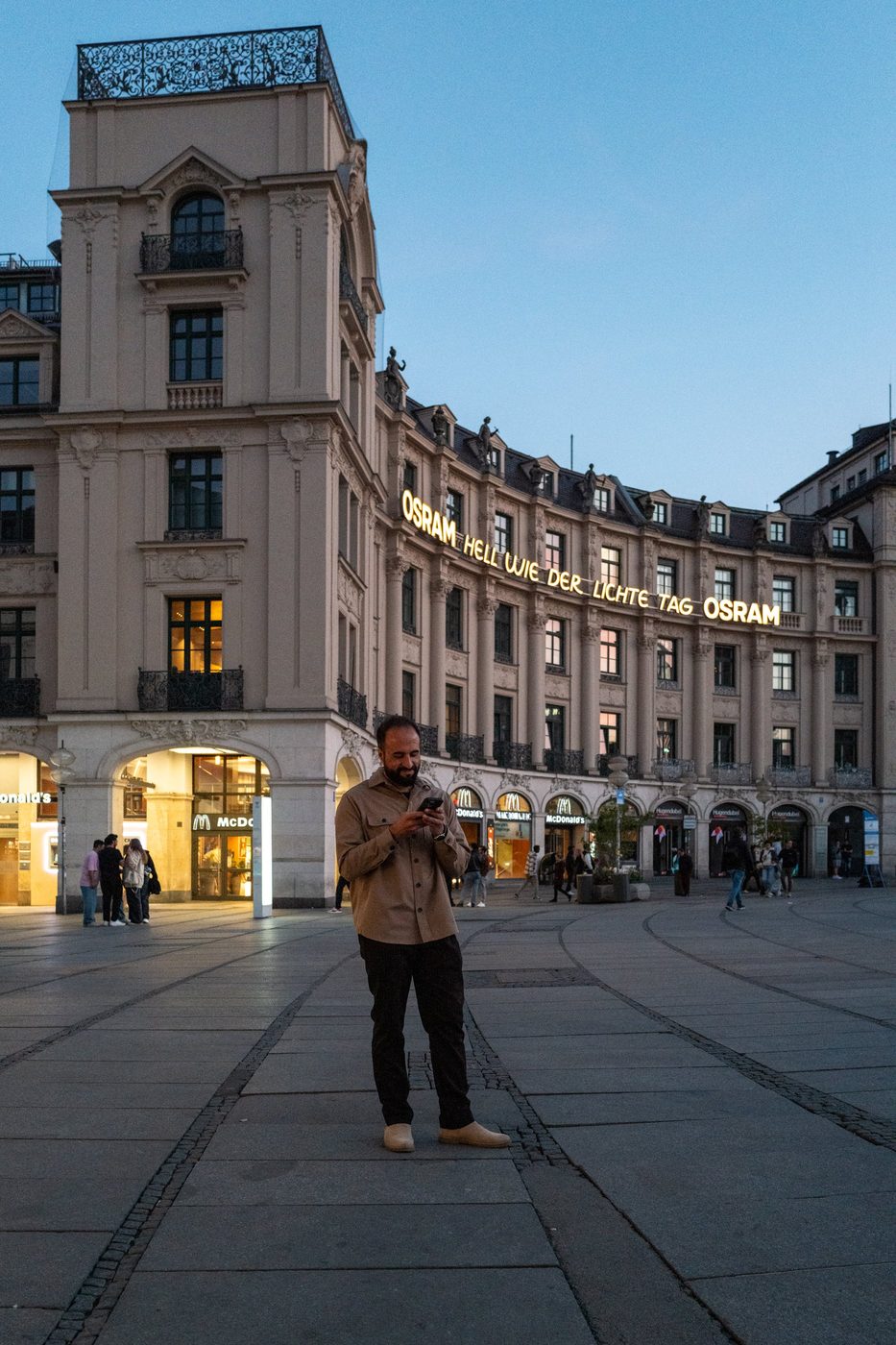 A man smiles while checking his phone in front of the illuminated Osram sign, Munich