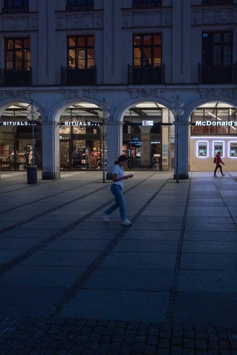 A woman walks alone at night under the illuminated arcades of a lively square, Munich