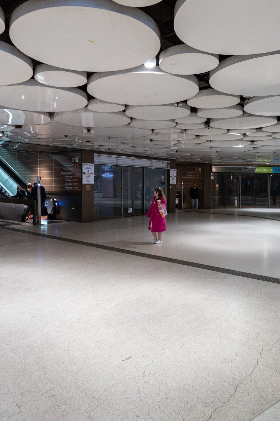 A passer-by in a pink coat crosses a station hall with an undulating ceiling, Munich