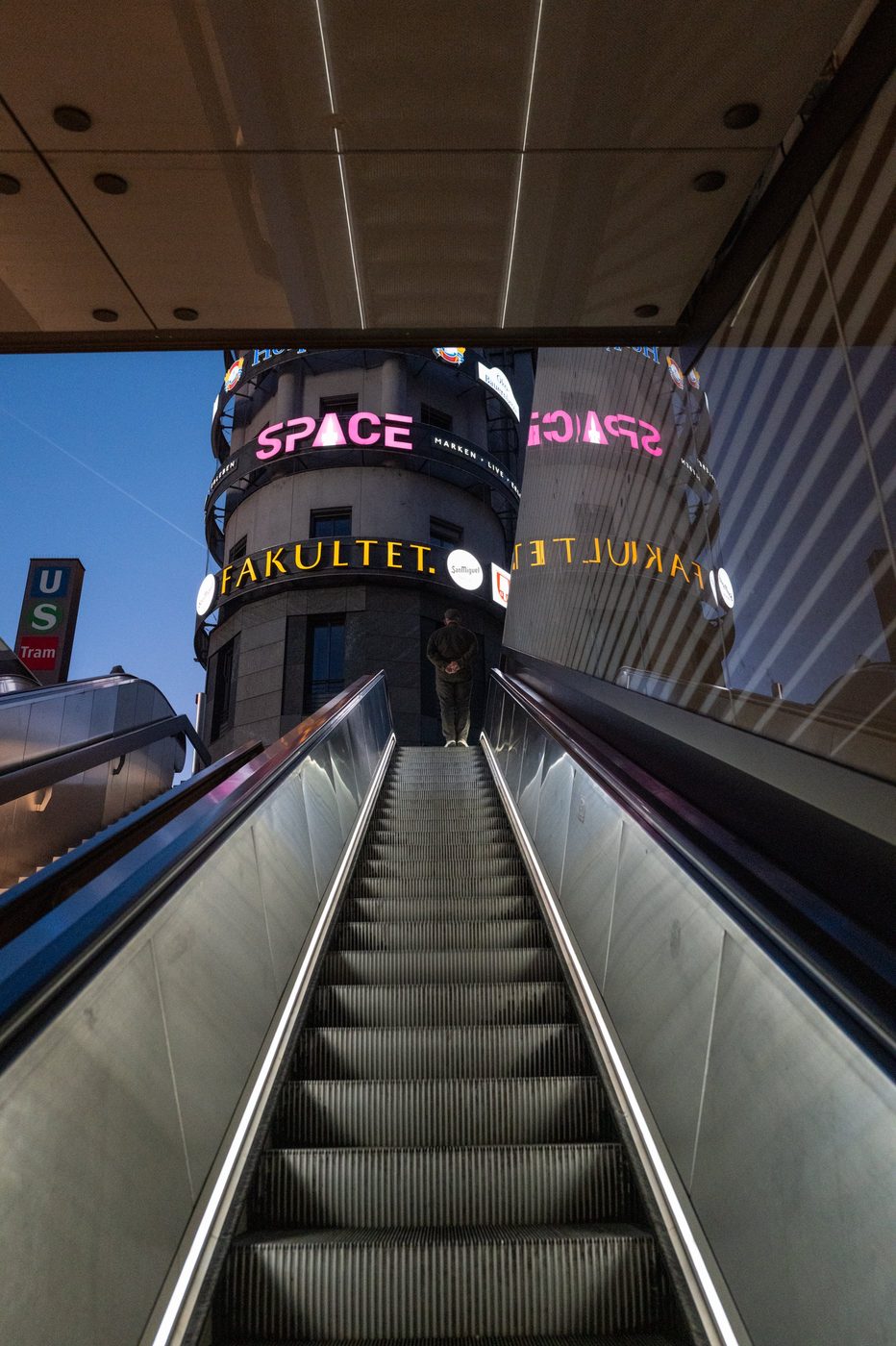 A man waits at the top of an escalator beneath the overlapping illuminated signs of a building, Munich