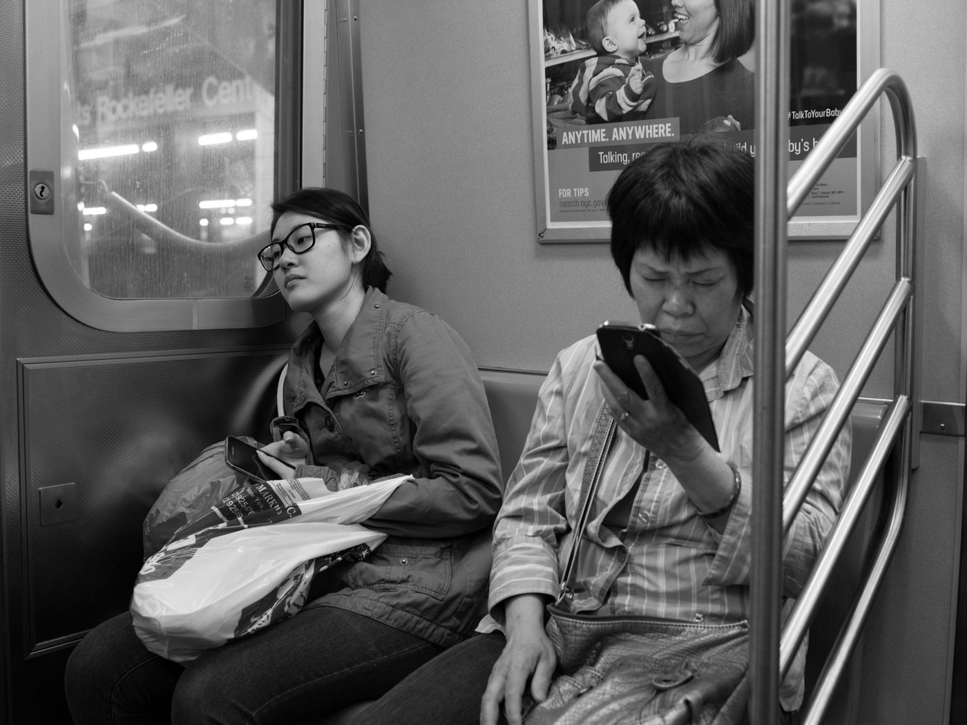 Two female subway passengers, one dozing against the window, the other on her phone, New York