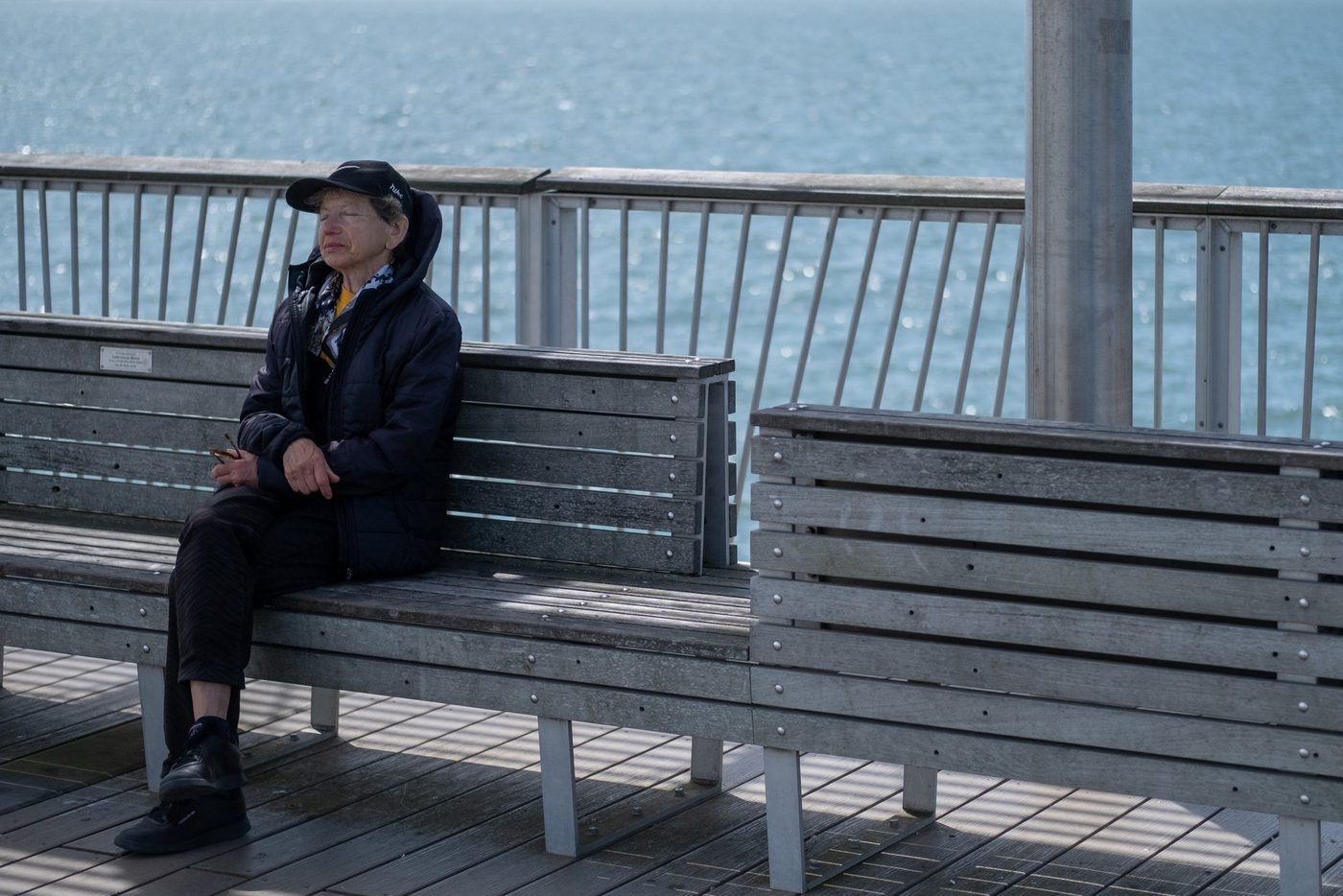 Elderly woman in a cap sitting alone on a bench facing the sea, New York