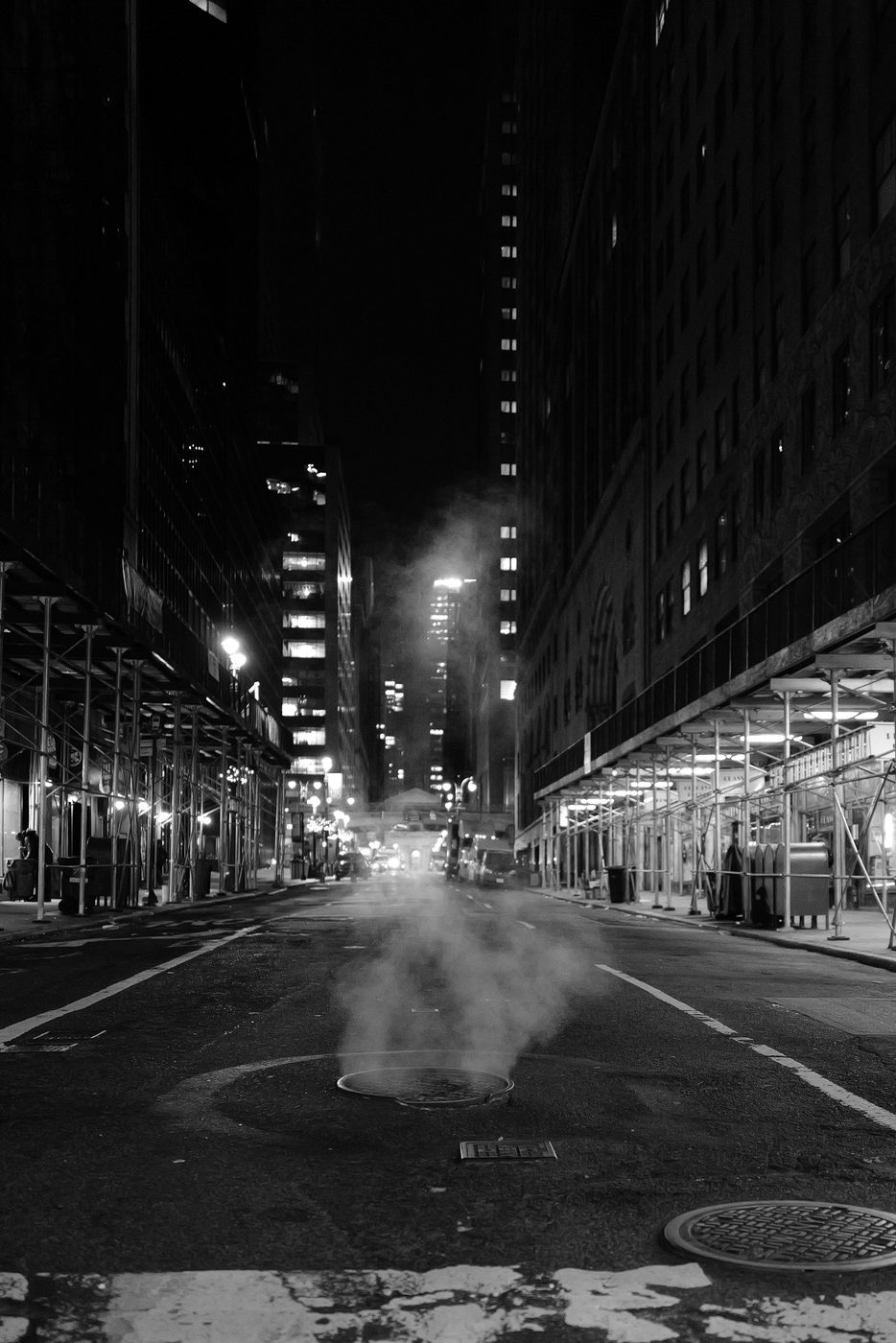 Steam rising from a manhole in the middle of a street lined with scaffolding, New York