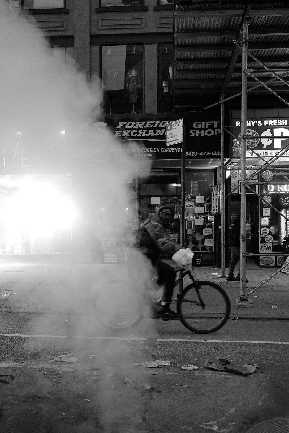 Cyclist crossing a cloud of steam in front of a currency exchange at night, New York