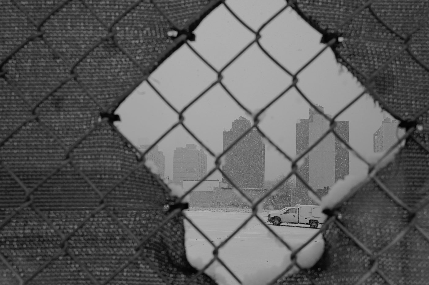 View of a snow-covered parking lot through a hole in a chain-link fence, New York