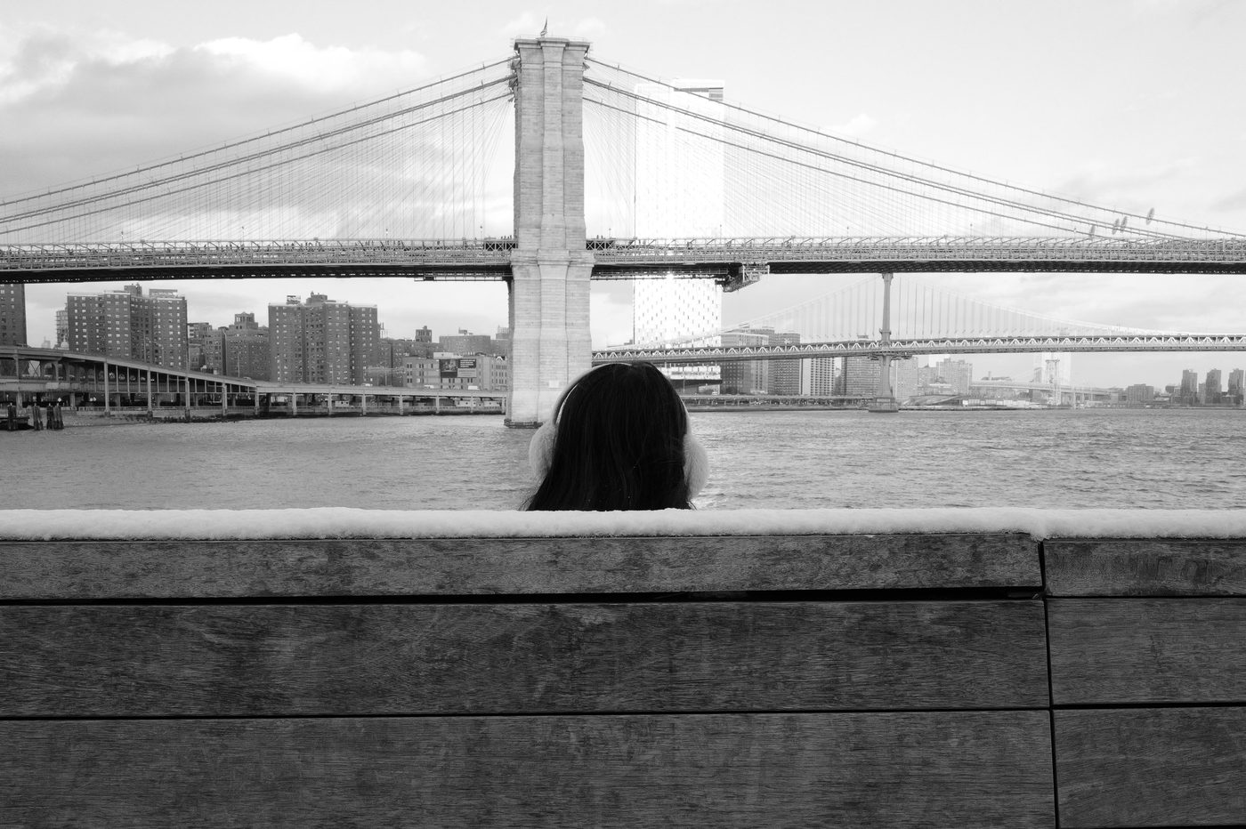 Woman in earmuffs contemplating the Brooklyn Bridge from a low wall, New York