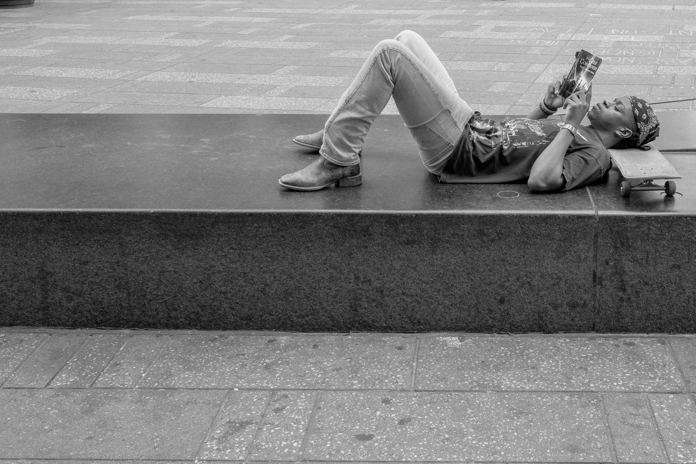 Young man lying on a low wall reading, skateboard set beside him, New York