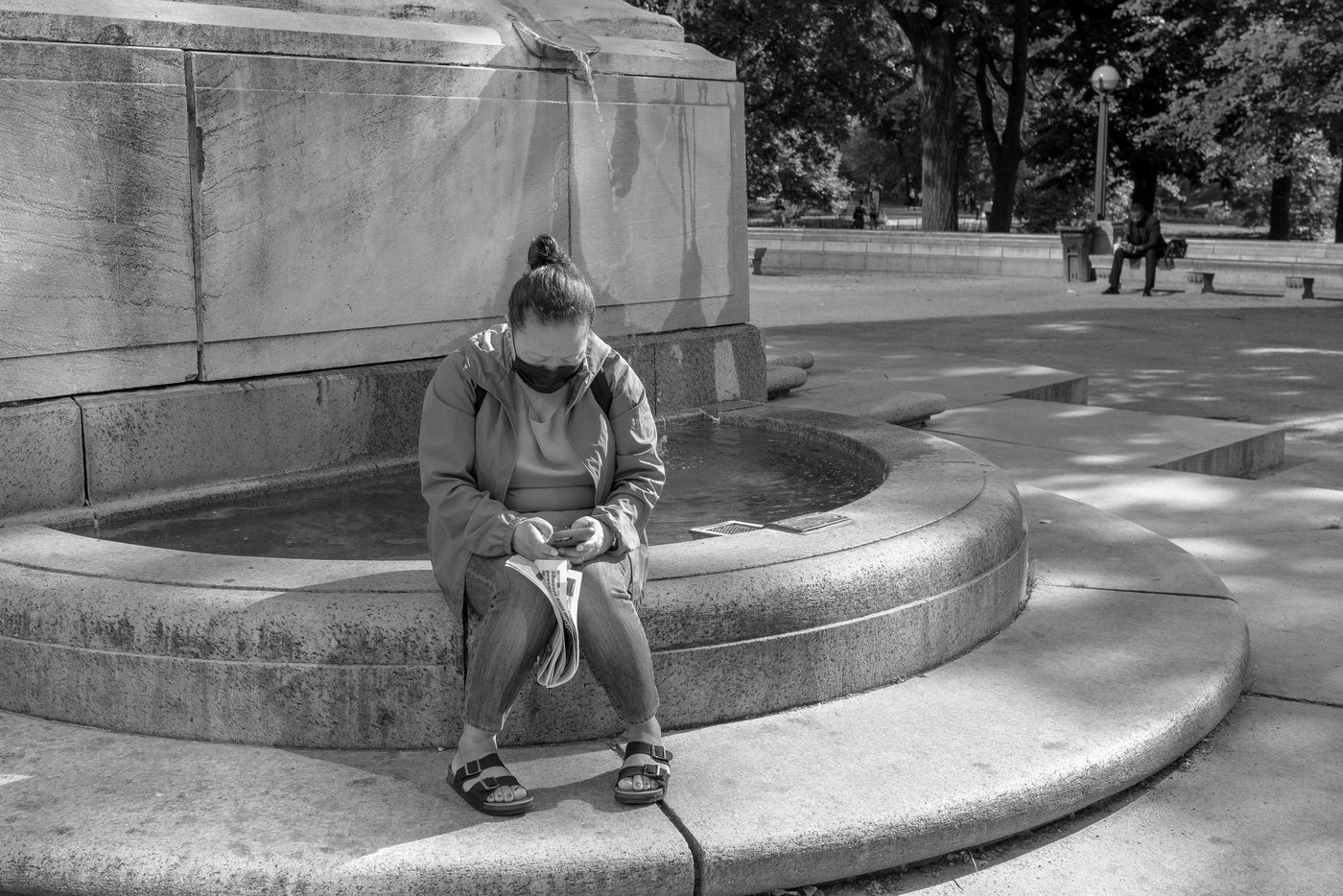Masked woman sitting at the edge of a fountain with her phone and a newspaper, New York