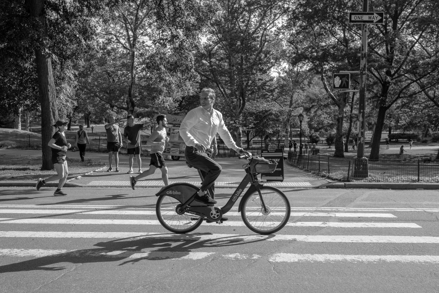 Man in a shirt pedaling a Citi Bike, joggers in the background, New York