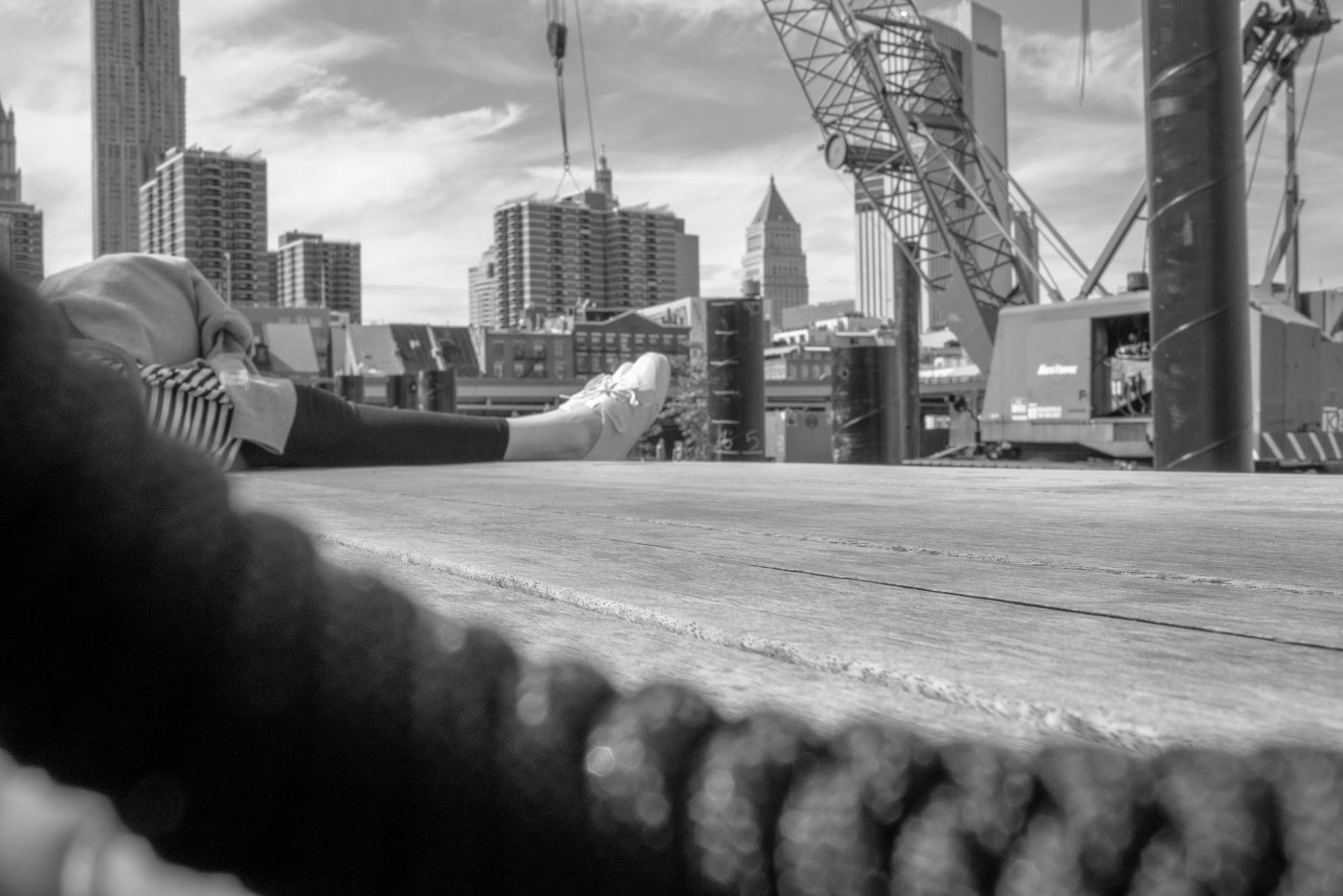 Feet resting on a wooden pier facing the skyline and a crane, New York