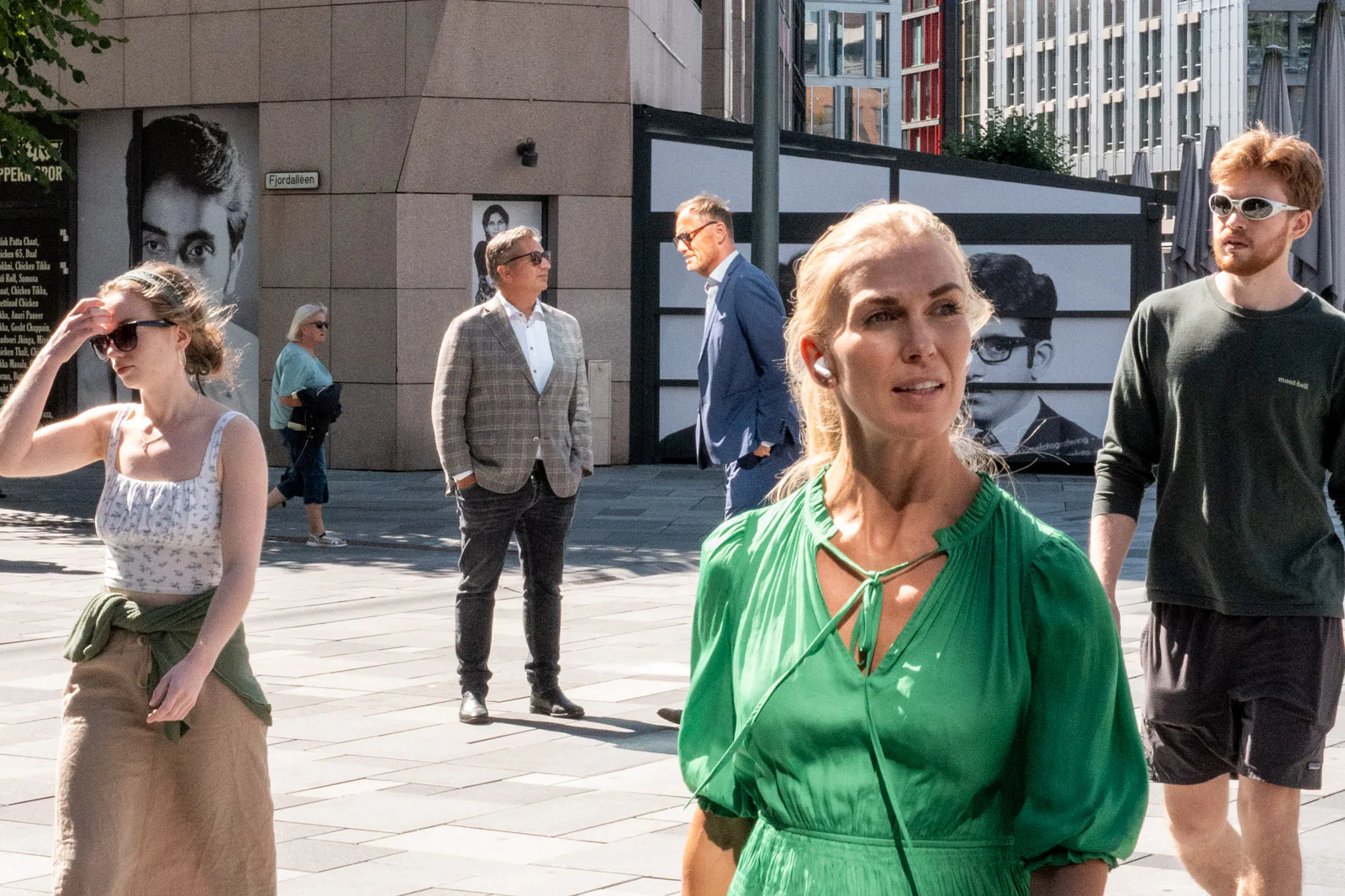 Des passants devant de grands portraits en noir et blanc, une femme en robe verte au premier plan, Oslo