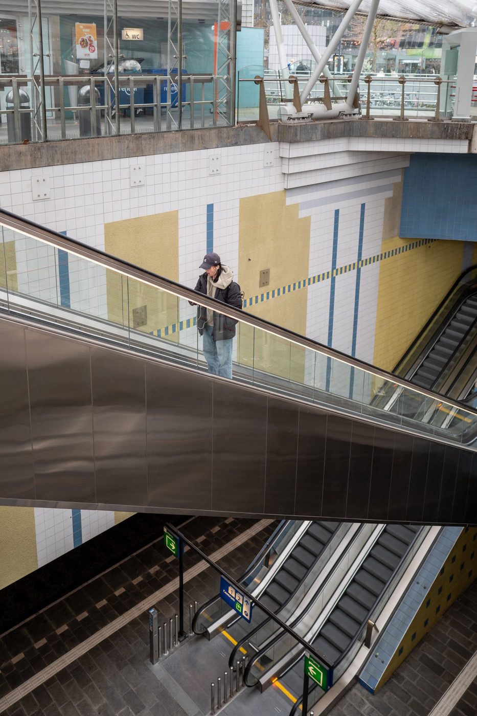 Passenger on a diagonal escalator in front of a yellow and blue tiled wall, Rotterdam