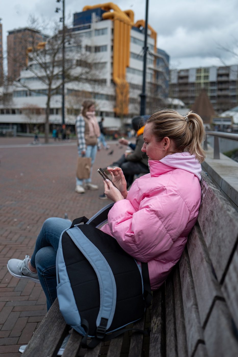 Young woman in a pink puffer jacket sitting on a bench checking her phone, Rotterdam