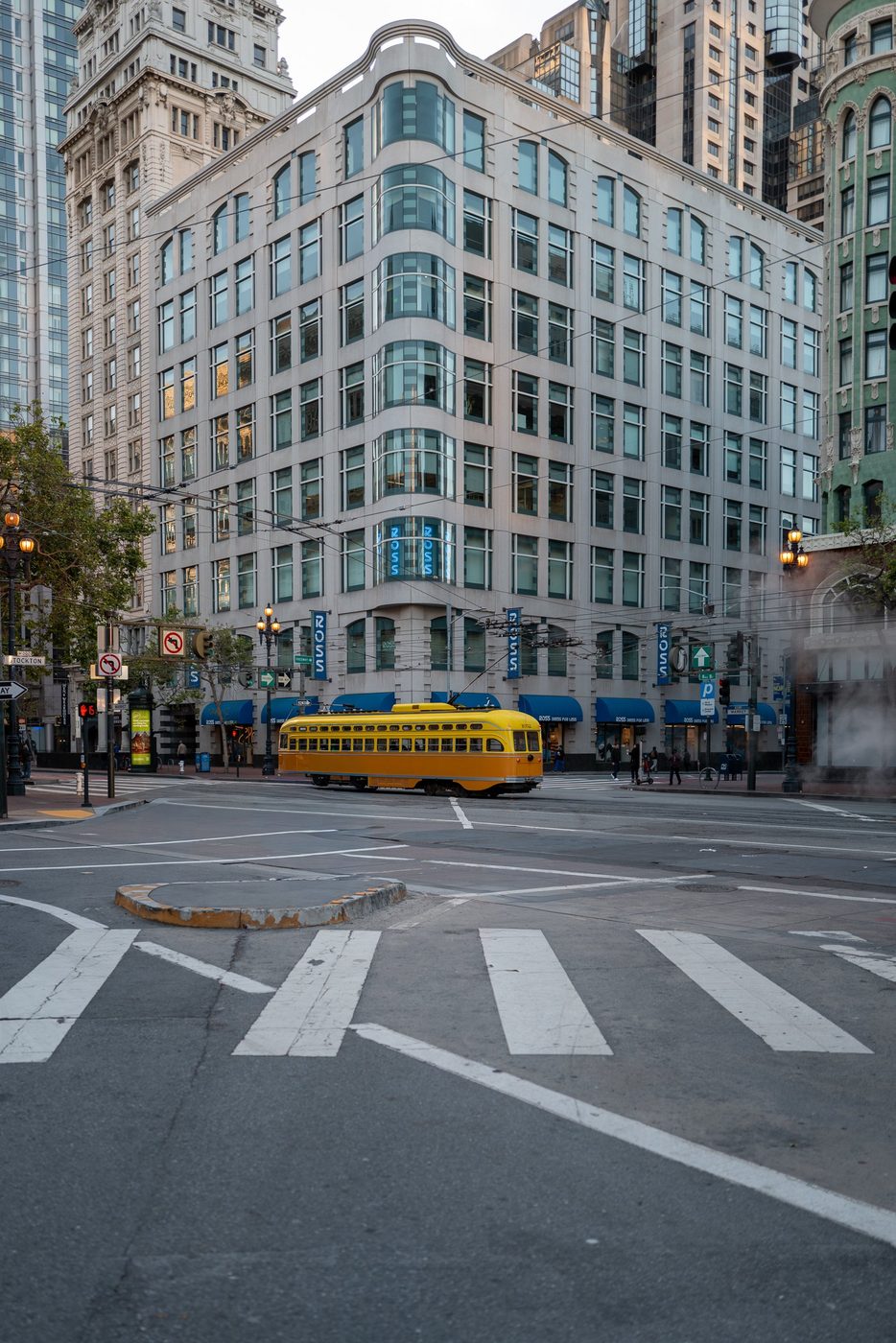 Historic yellow streetcar crossing a deserted intersection in front of the curved façade of the Ross store, San Francisco