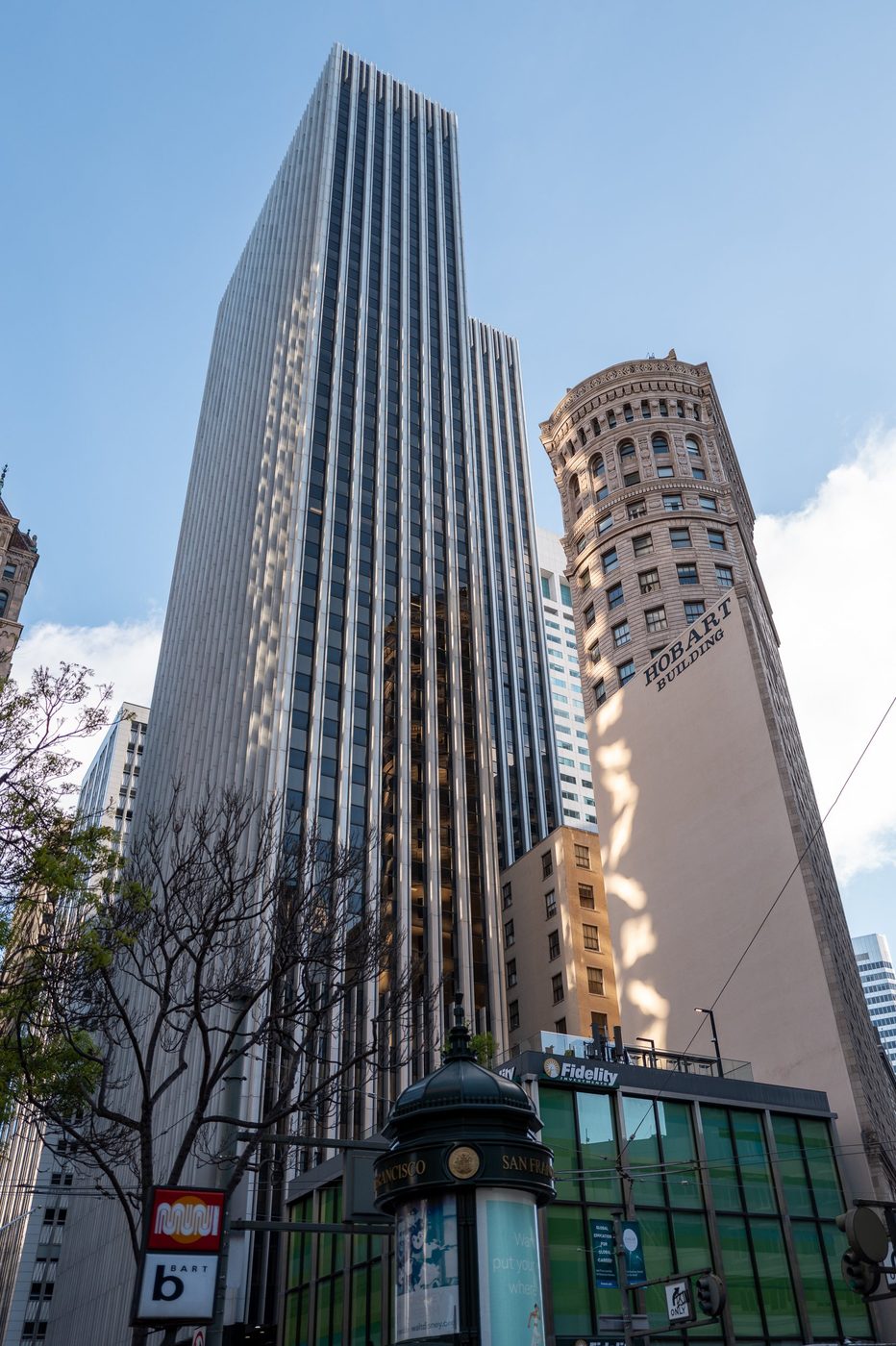 Low-angle view of the Hobart Building and a neighboring skyscraper, Muni and BART signs on the ground, San Francisco