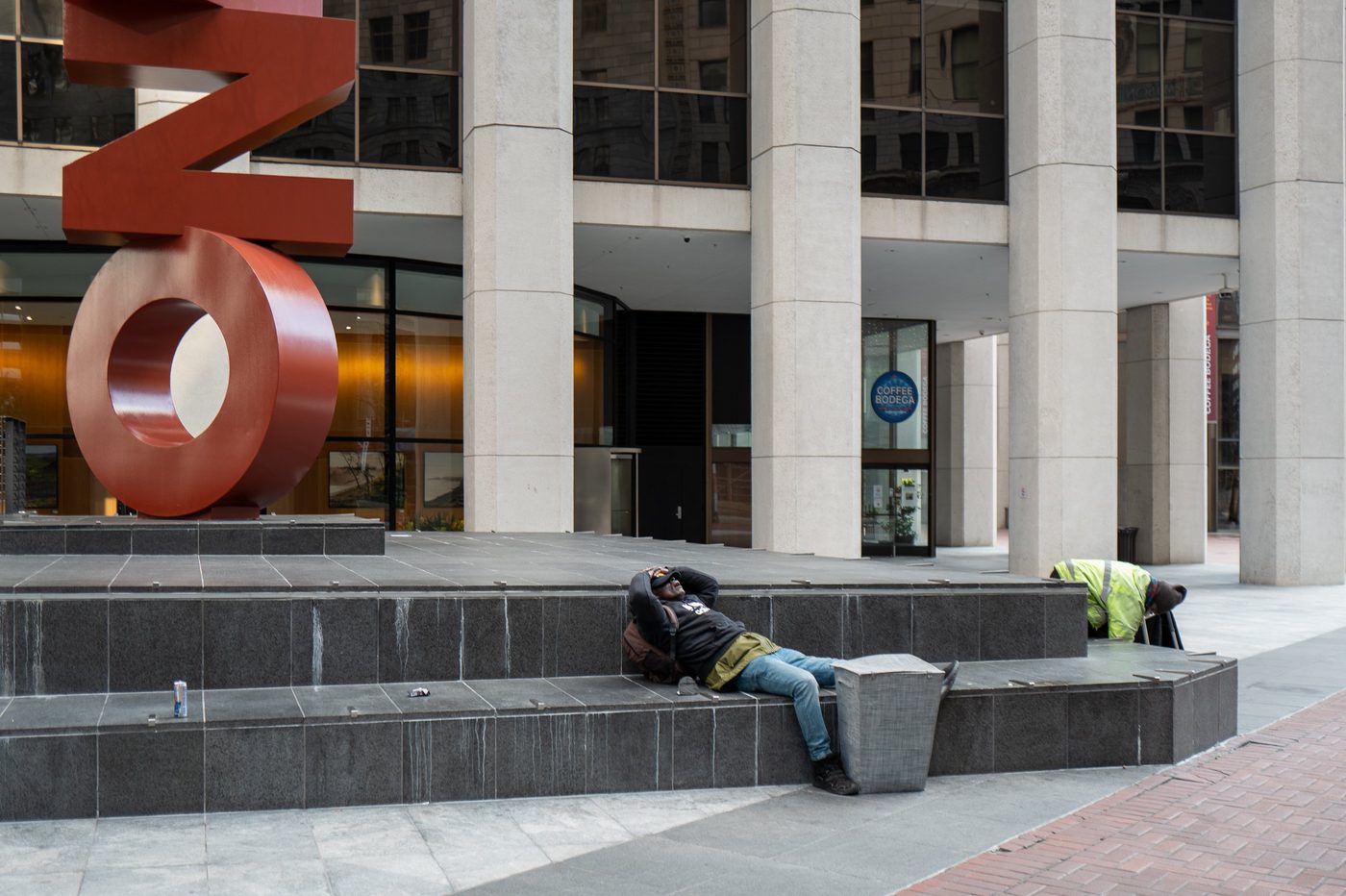 Man lying down resting on a granite bench near a monumental red sculpture, San Francisco
