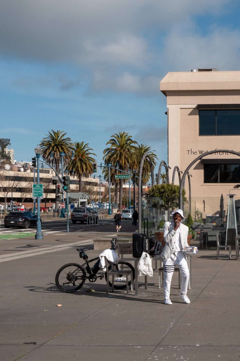 Man in a white suit sitting near his bike and suitcases, palm trees in the background, San Francisco