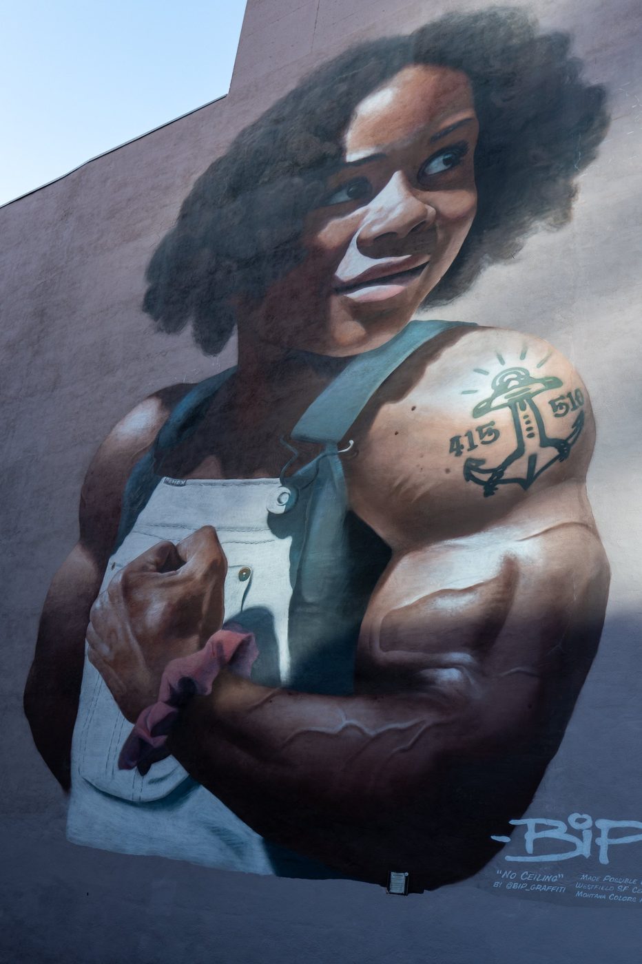 Mural of a muscular woman in overalls, arm raised and tattoo on her shoulder, San Francisco