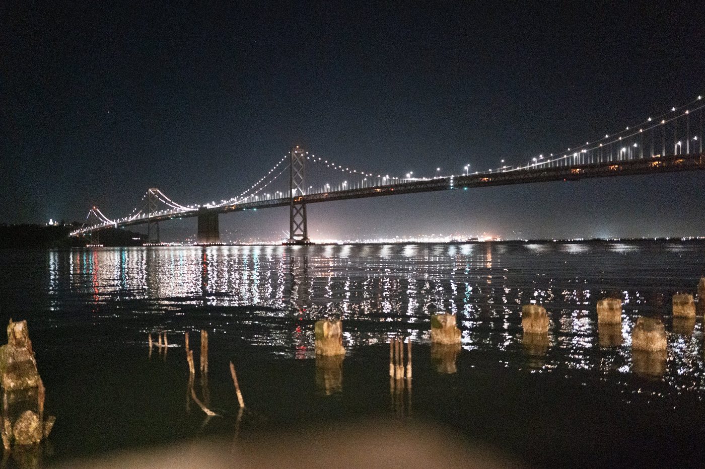 Illuminated Bay Bridge reflecting on the dark water, pilings in the foreground, San Francisco