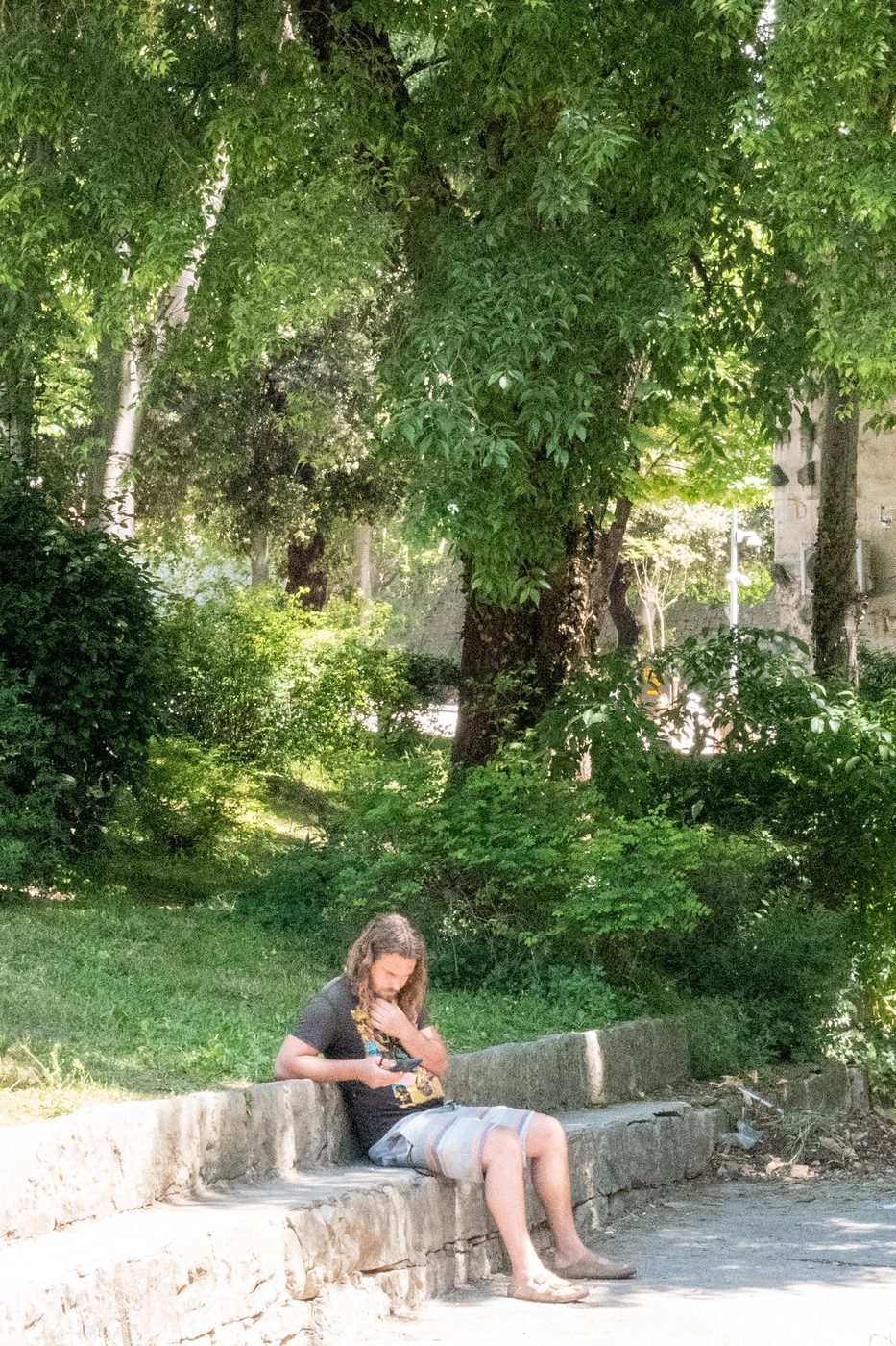 Man sitting on a low wall checking his phone in the shade of the trees, Split
