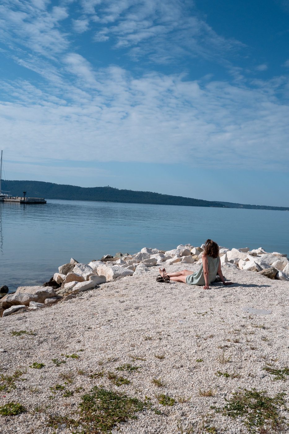 A passerby sitting on the rocks facing the calm bay, Trogir
