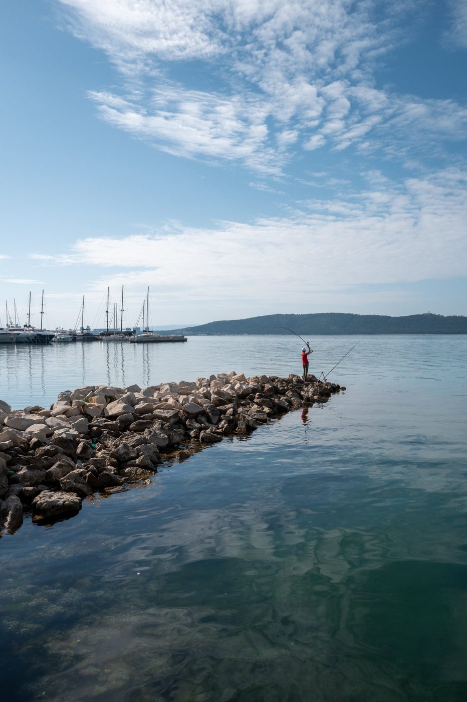 Fisherman mid-cast on the jetty, sailboats in the background, Trogir