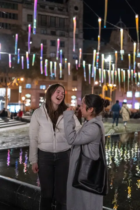Deux jeunes femmes riant devant un bassin reflétant des guirlandes lumineuses, Barcelone