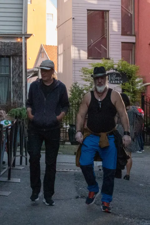 Two men walking down the street, one in a tank top and hat, Bergen