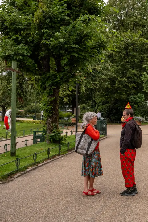 A lady in a flowered dress and a man in red trousers chatting in a park, Bergen