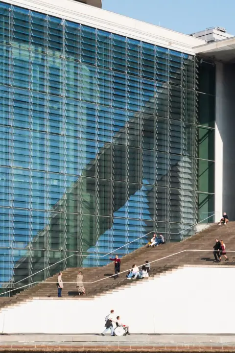 Reflets diagonaux sur une façade de verre, passants assis sur un escalier, Berlin
