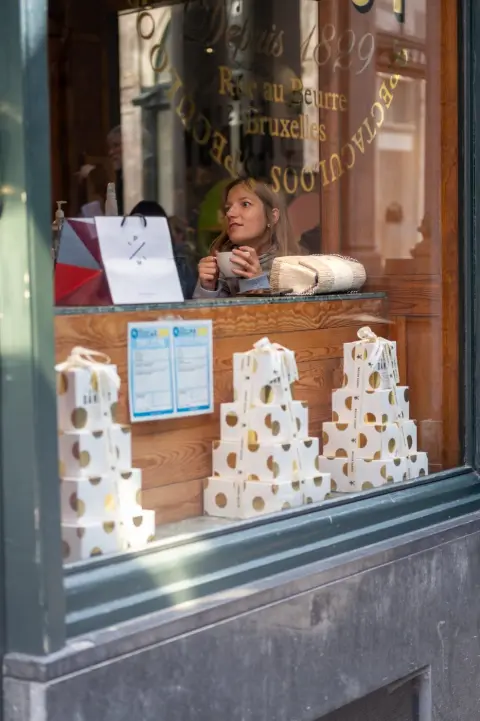 Vendeuse derrière la vitrine d’une chocolaterie, boîtes à pois dorés empilées au premier plan, Bruxelles