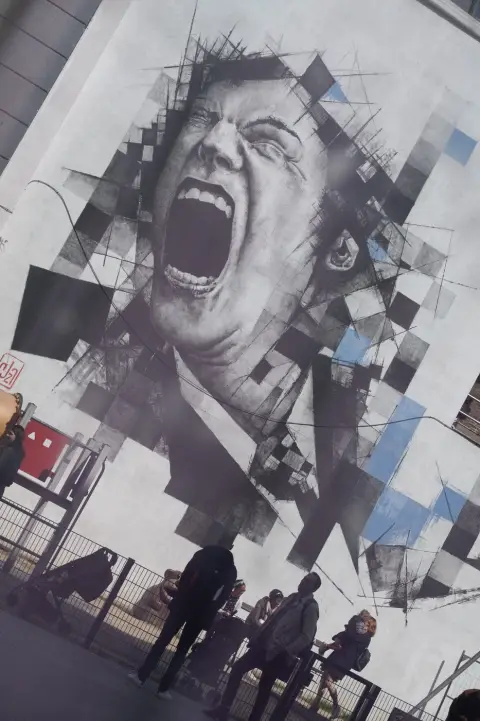 Fresque d’un visage qui crie, éclaté en carrés, au-dessus de passants à contre-jour, Bruxelles