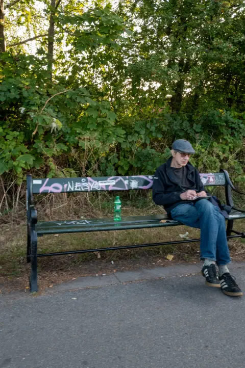 A man in a cap sitting on a graffitied bench at the edge of a wood, Copenhagen
