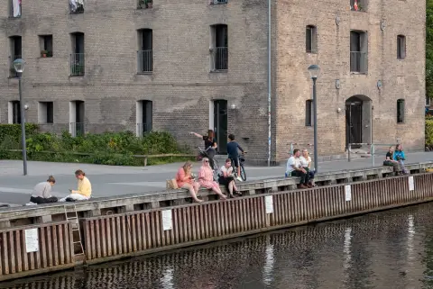 Young people sitting on the edge of a quay in front of a brick warehouse, Copenhagen