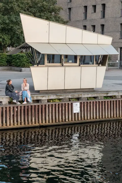 Two women sitting on the edge of the quay near a kiosk with folded roofs, Copenhagen