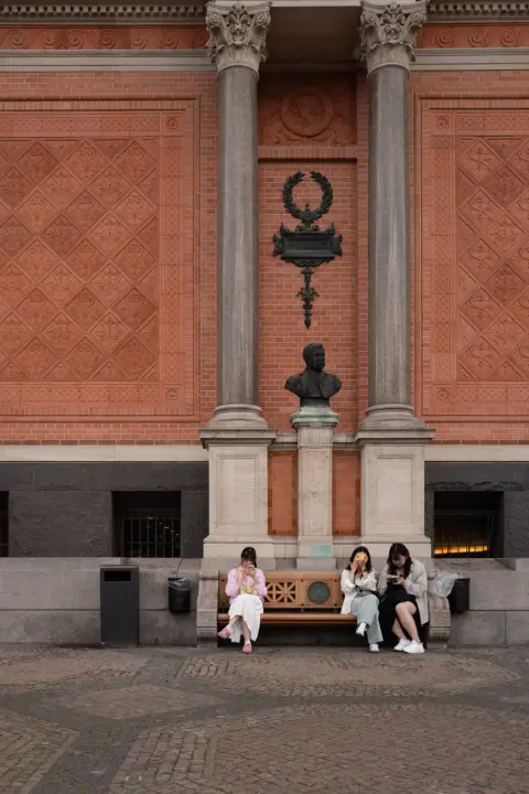Three young women on a bench beneath a bust, in a red brick niche, Copenhagen