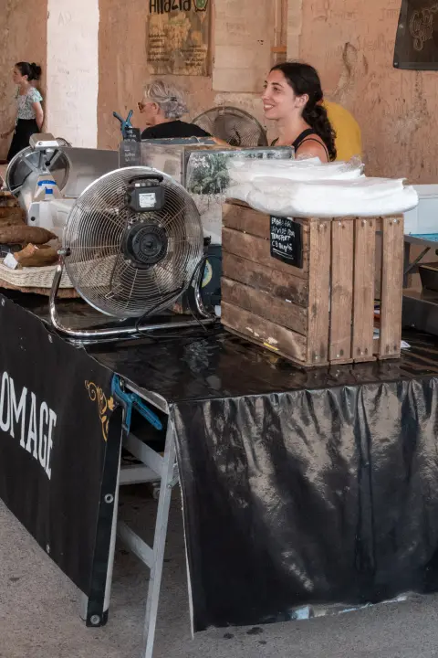Une fromagère sourit derrière son étal, un ventilateur au premier plan, Bonifacio, Corse