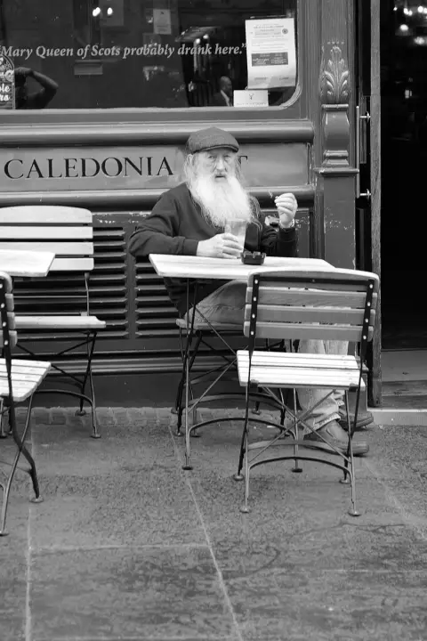 Old man with a white beard at a table beneath a tongue-in-cheek sign about Mary Queen of Scots, Edinburgh