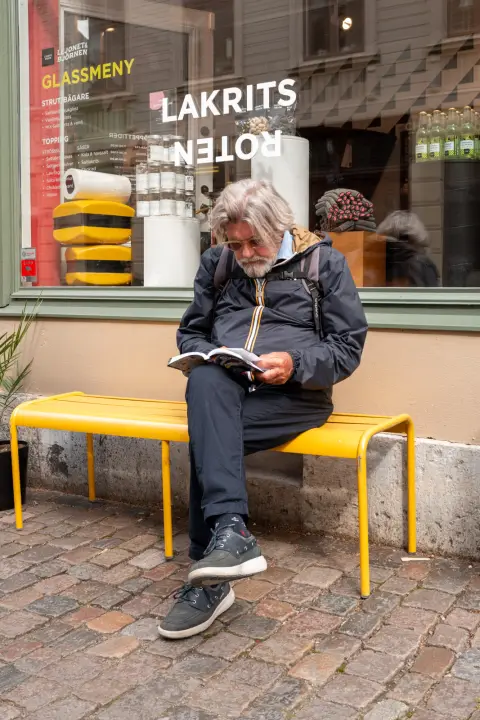 A white-bearded man reading on a yellow bench in front of a liquorice shop, Gothenburg