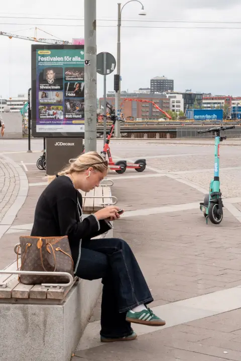 A woman checking her phone on a bench, scooters behind her, Gothenburg