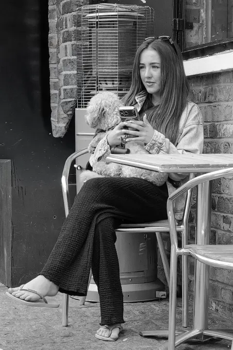 Jeune femme en terrasse tenant son petit chien tout en regardant son téléphone, Londres