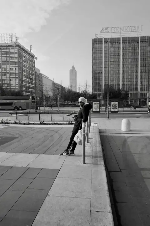 Jeune femme au foulard adossée à une barrière, téléphone en main, tours modernes en arrière-plan, Milan