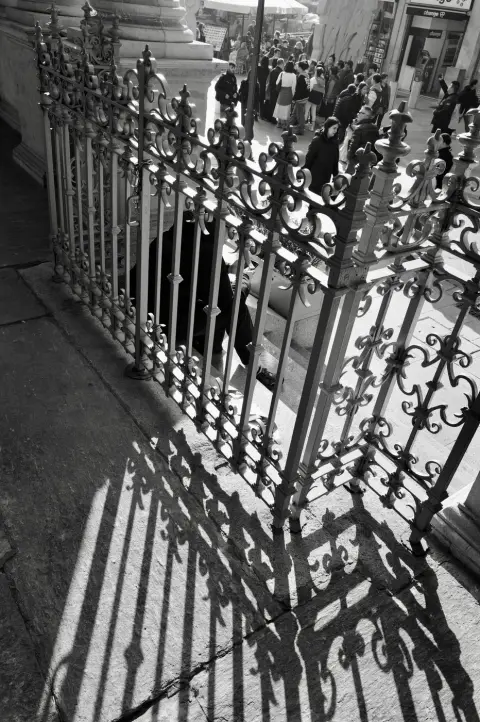 Grille en fer forgé projetant ses ombres sur le pavé, foule floue à l'arrière-plan, Milan