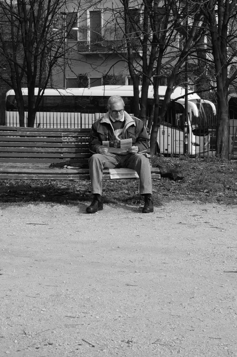 Homme âgé lisant son journal sur un banc public, devant un autocar garé, Milan