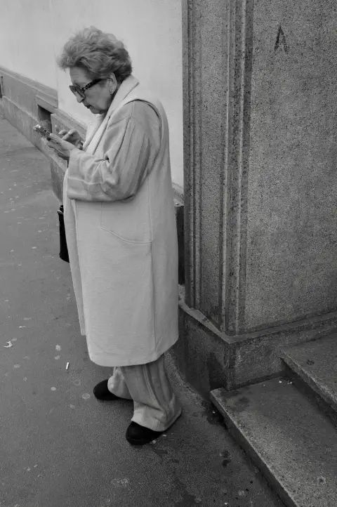 Femme âgée en manteau clair consultant son téléphone, adossée à un mur de pierre, Milan