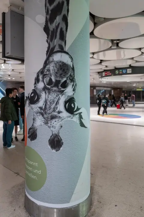 Affiche publicitaire d'un animal accroché la tête en bas sur un pilier de gare, Munich